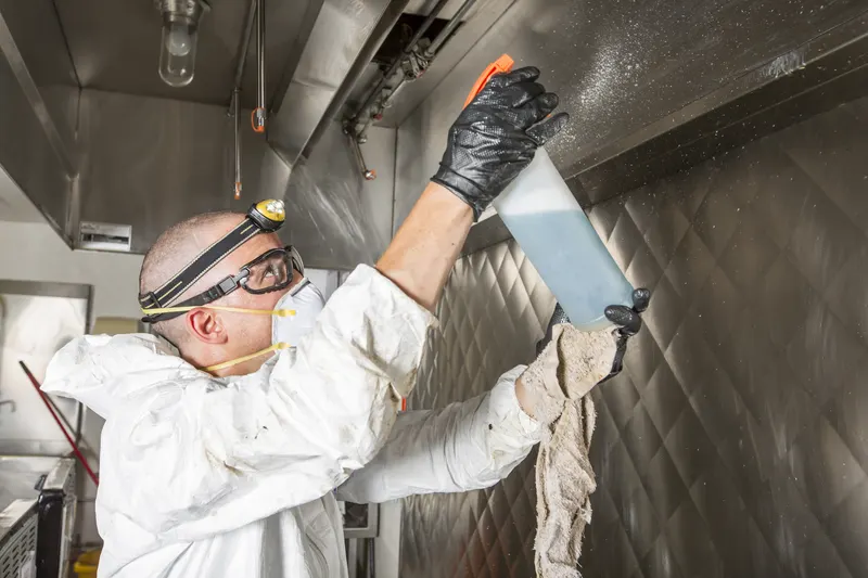 Technician spraying degreaser overhead inside a commercial kitchen exhaust hood with a cleaning cloth in the other hand
