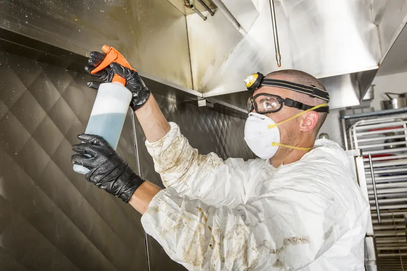 Technician in protective coveralls spraying degreaser on a stainless steel kitchen wall below an exhaust canopy