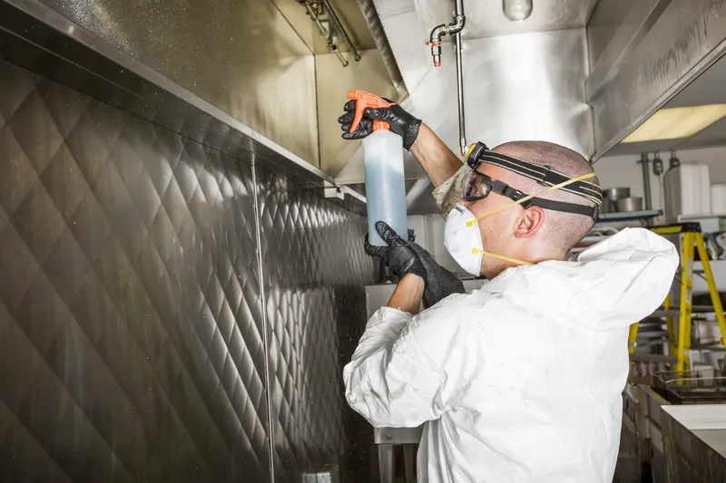 Technician in coveralls spraying degreaser onto the inside face of a stainless steel exhaust canopy with the baffle filters removed
