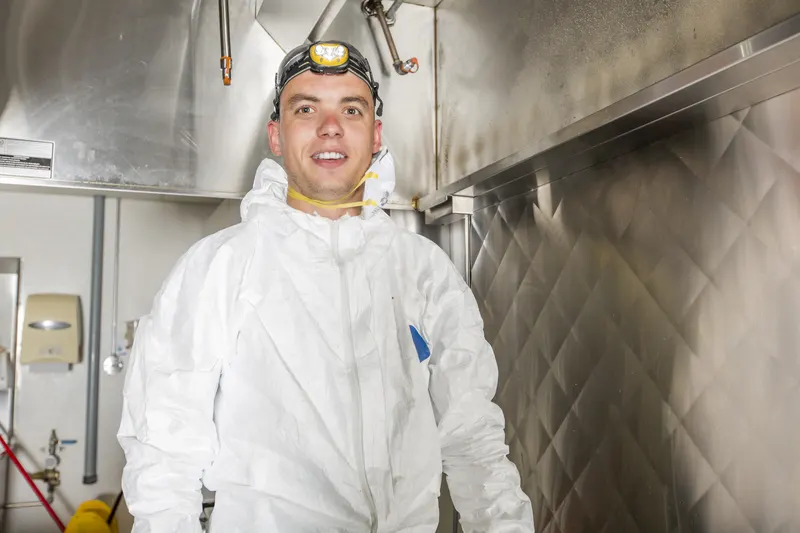 Hood cleaning technician in white coveralls and a headlamp standing under a stainless steel canopy in a commercial kitchen