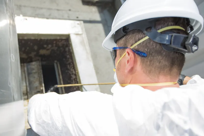 Technician in a white hard hat and safety glasses reaching into an open access hatch on a grease-coated commercial kitchen exhaust duct