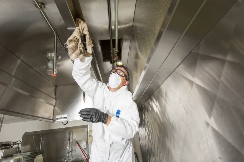 Gloved hand wiping the plenum wall above the filter bank inside a commercial kitchen exhaust hood