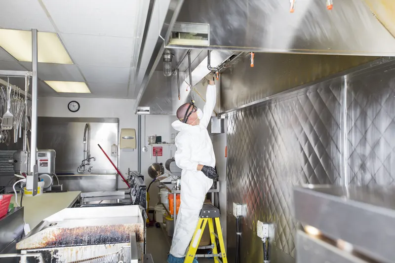 Hood cleaning technician on a step ladder cleaning a stainless steel canopy in a full commercial kitchen with the cook line covered