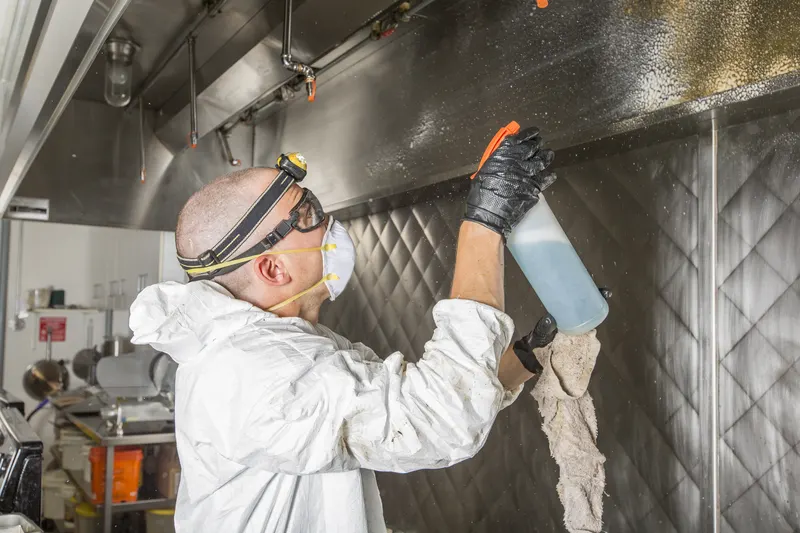 Technician in white coveralls, safety goggles and a headlamp wiping the underside of a stainless steel canopy hood with a cloth while holding a spray bottle over a commercial cook line